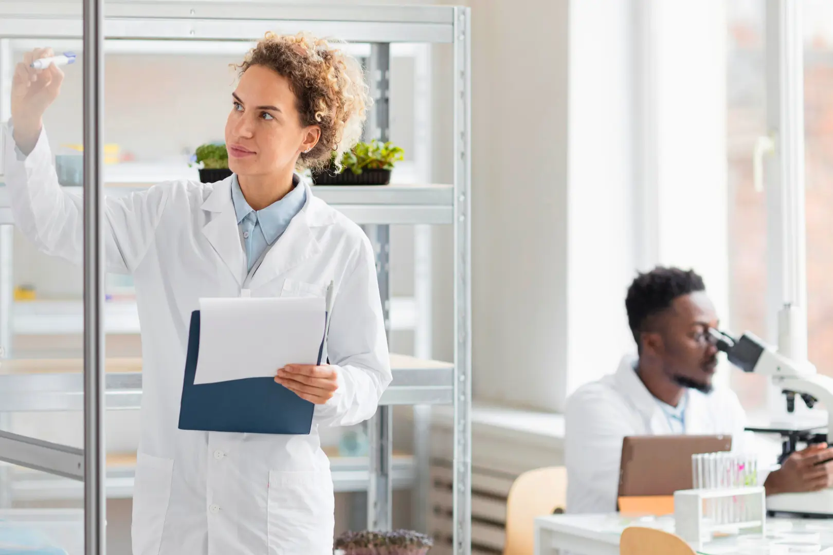 An Abuse Counseling & Therapy researcher writes on a glass board while a colleague uses a microscope.