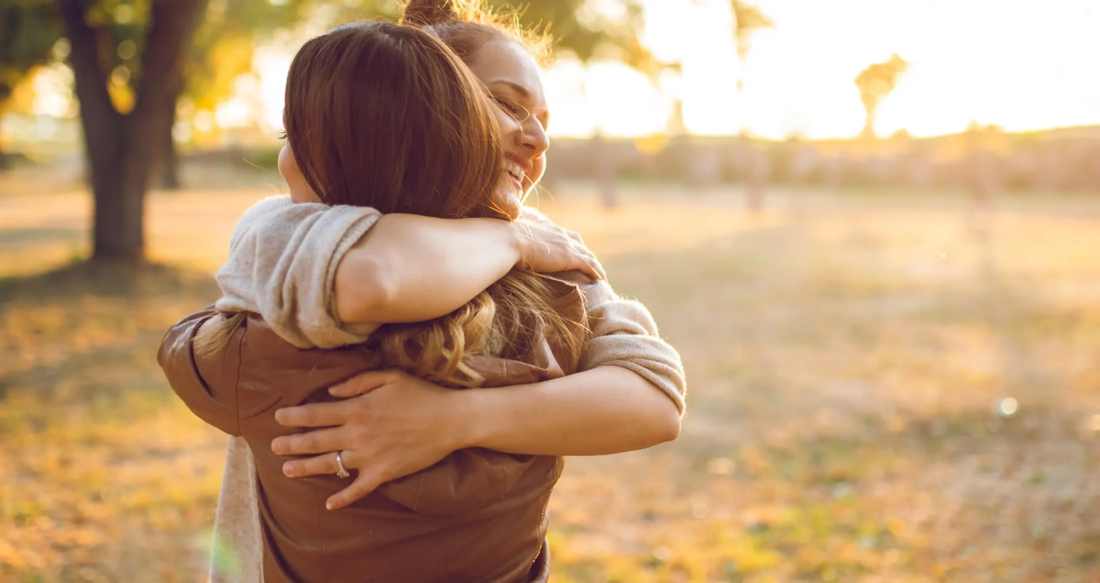 Two women share a warm, supportive embrace in a sunny park, representing Abuse Counseling & Therapy.
