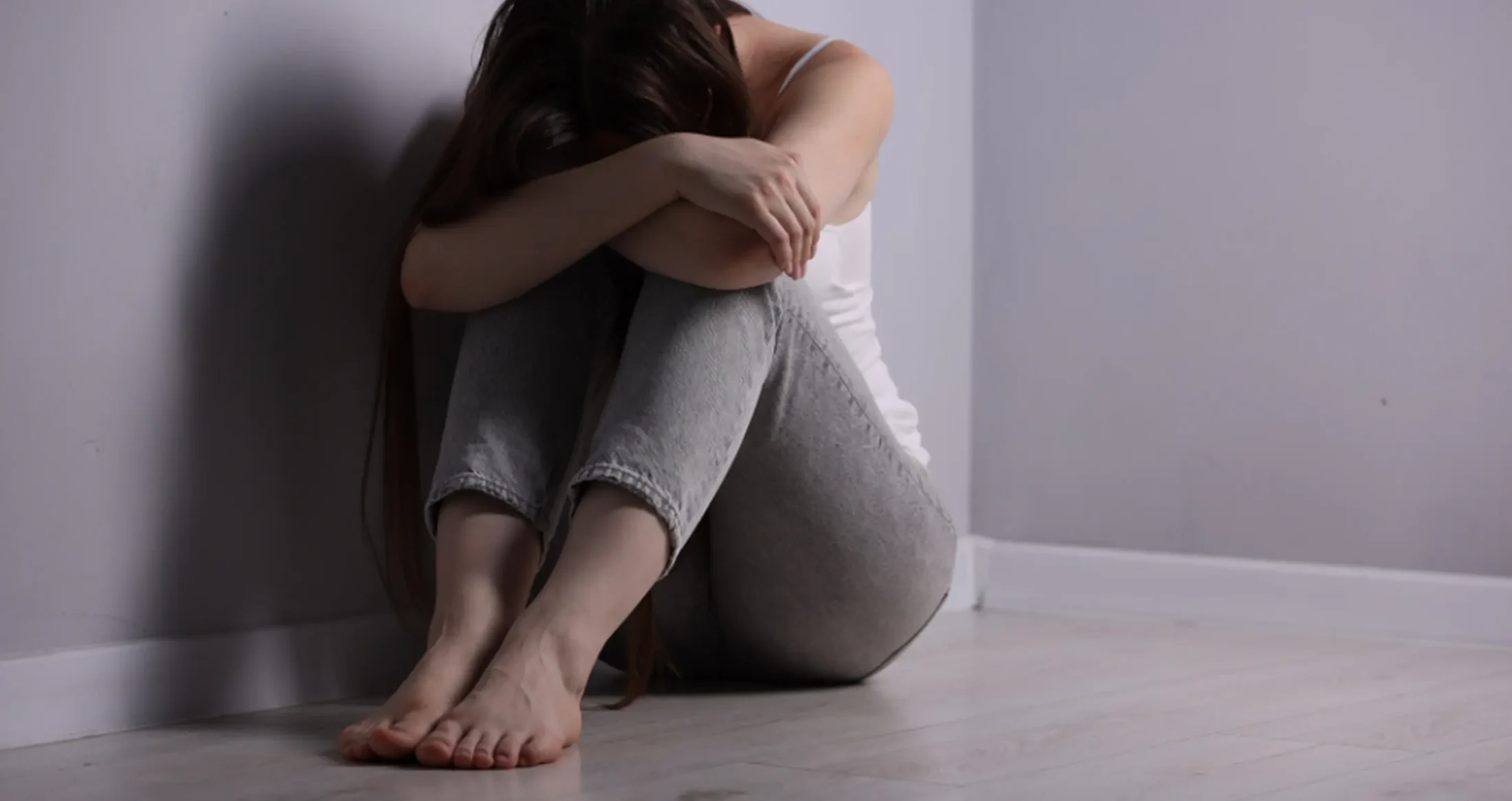 A woman sits curled on the floor in a corner, representing survivors helped by Abuse Counseling & Therapy.